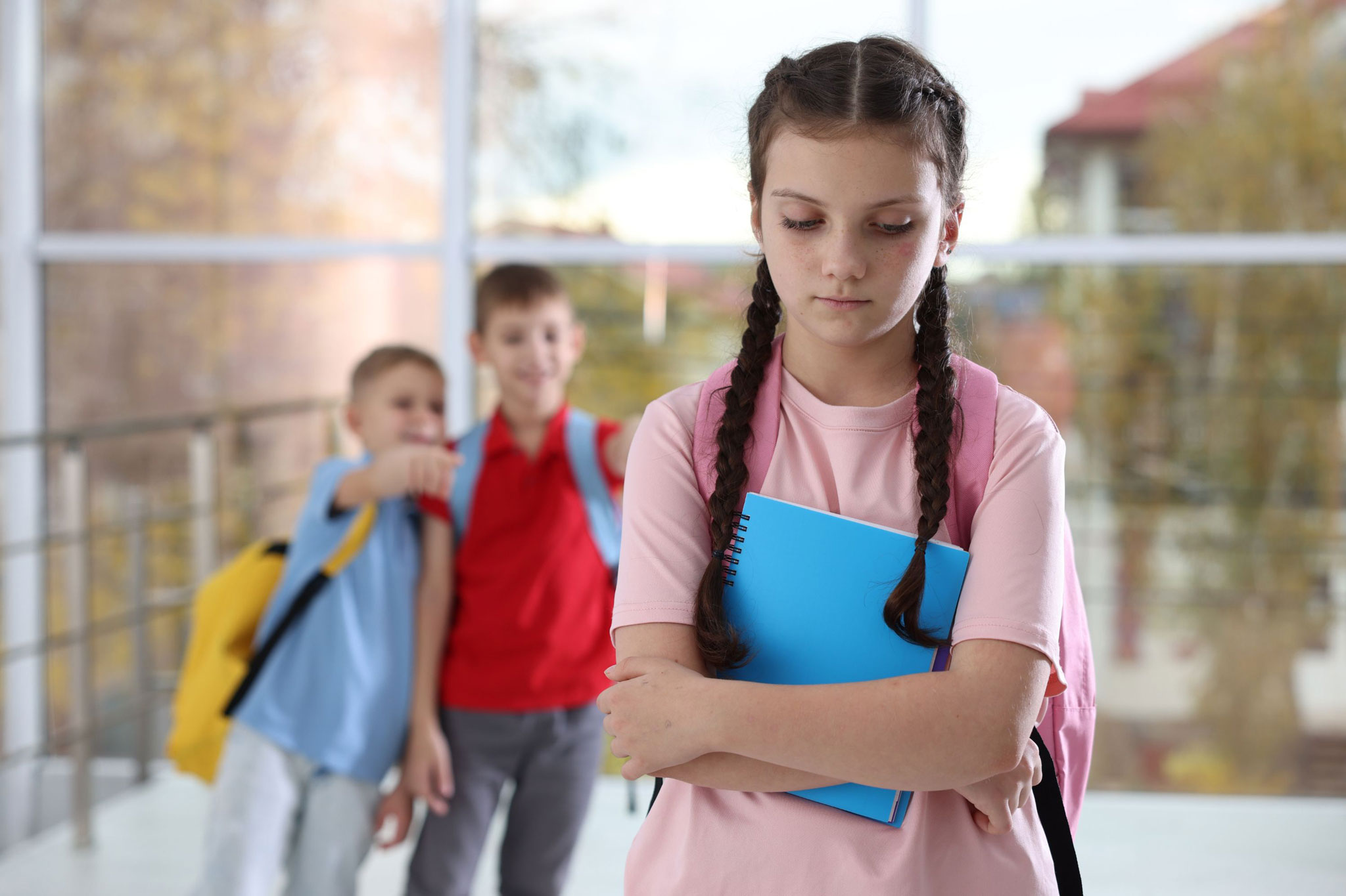 A young girl stands alone in a school hallway looking down with crossed arms while two boys behind her point and laugh, illustrating in-person bullying at school